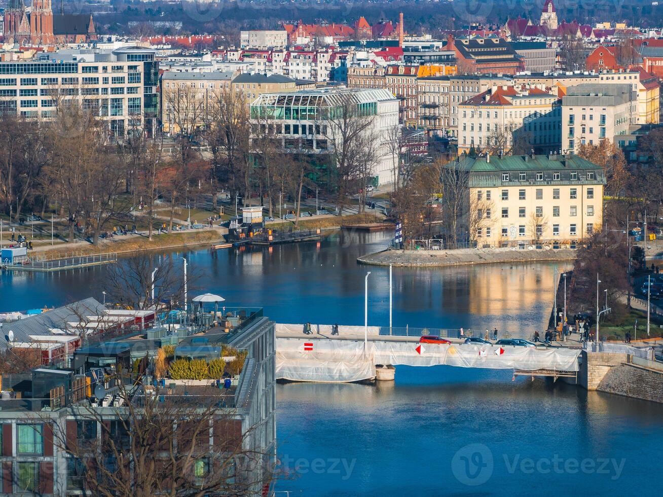Aerial view of Wroclaw, Poland, showing a river, a partially covered bridge, historic and modern buildings, a church spire, and leafless trees. photo