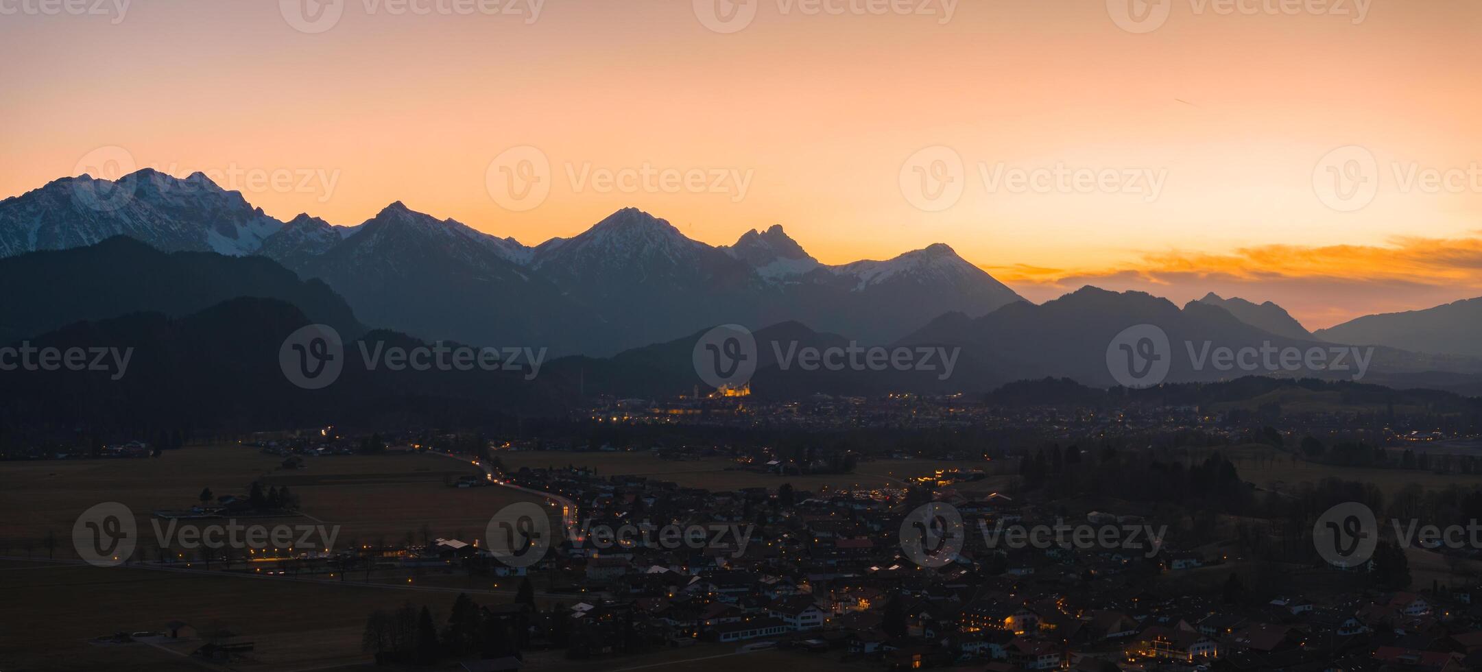Neuschwanstein Castle glows at sunset, surrounded by dark hills, the Bavarian Alps, and a village with scattered lights in the foreground. photo