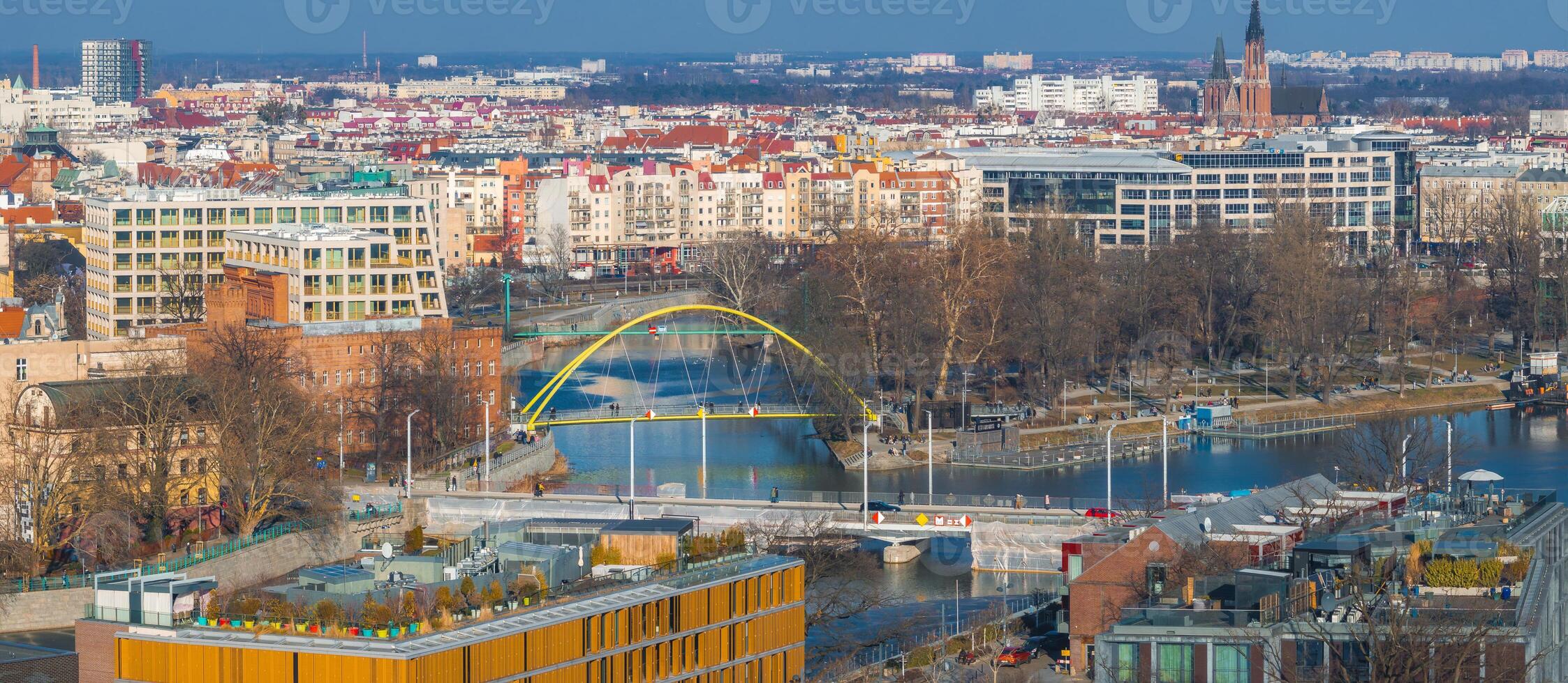 Aerial view of Wroclaw, Poland, showing a yellow arched pedestrian bridge, Gothic church tower, modern and historic buildings, parks, and a waterway. photo
