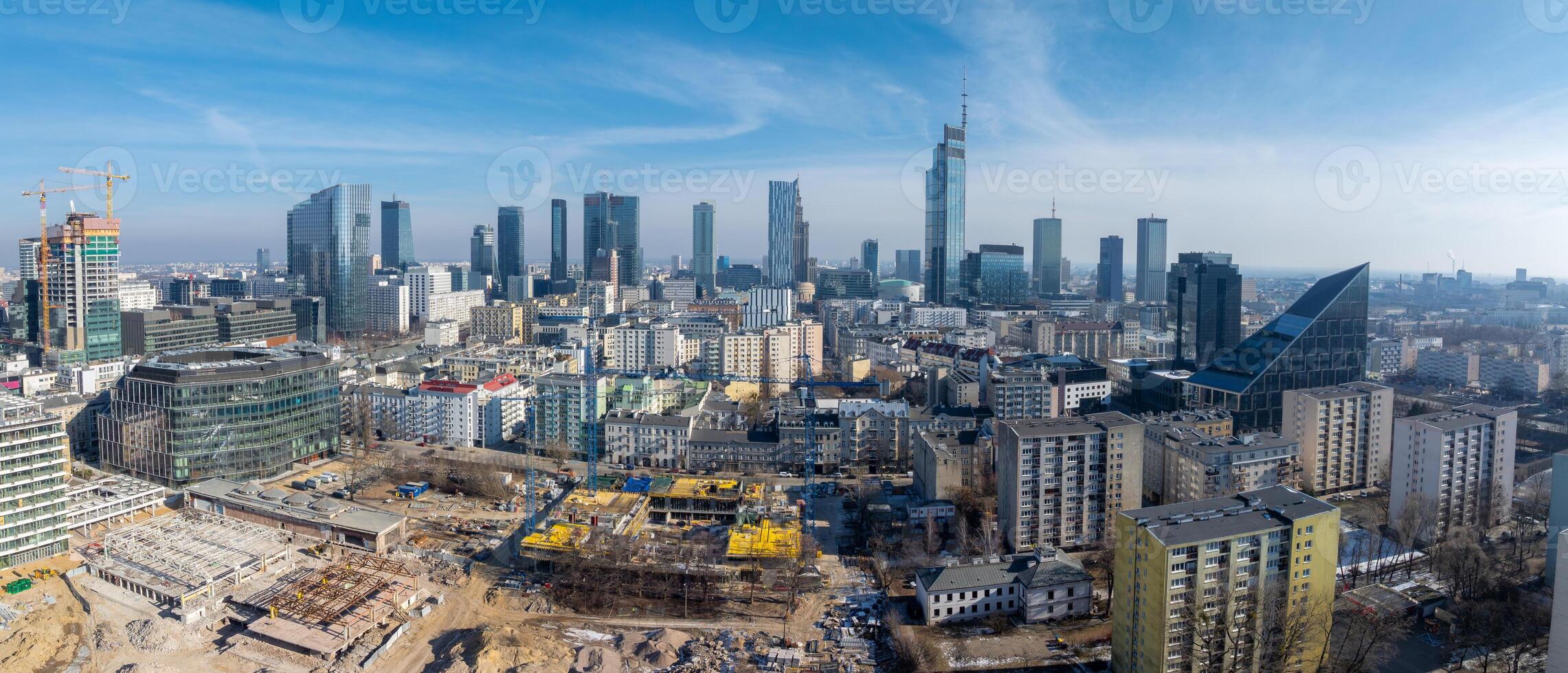Aerial view of Warsaw, Poland, featuring Varso Tower, construction sites, mid rise buildings, and a triangular glass roofed structure under a clear sky. photo