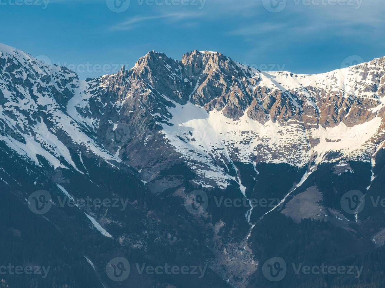 Snow capped peaks of the Alps near Innsbruck, Austria, with rugged ridges, rocky outcrops, forested slopes, and a clear blue sky in view. photo