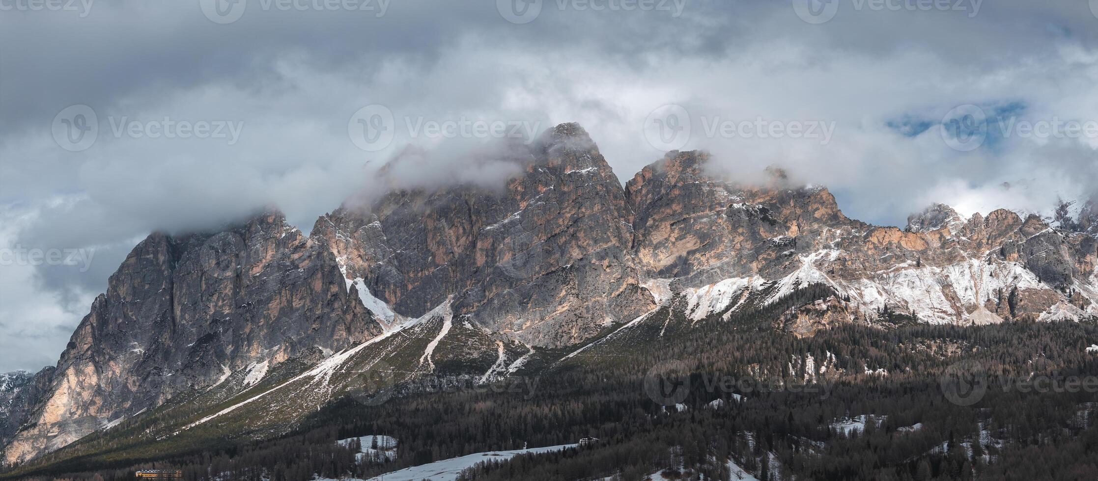 Cloud covered Dolomite peaks with snow patches rise above forests near Cortina d'Ampezzo, Italy, showing hints of human activity in the landscape. photo