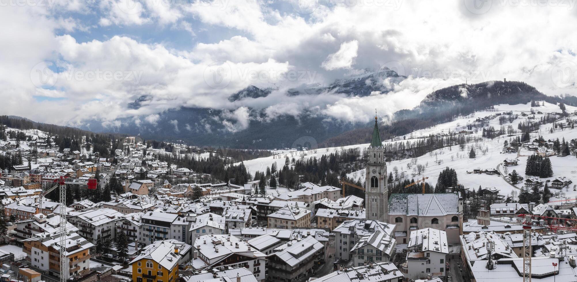 Snow covered rooftops and the Basilica Minore dei Santi Filippo e Giacomo stand out in Cortina d'Ampezzo, surrounded by Dolomites and construction cranes. photo