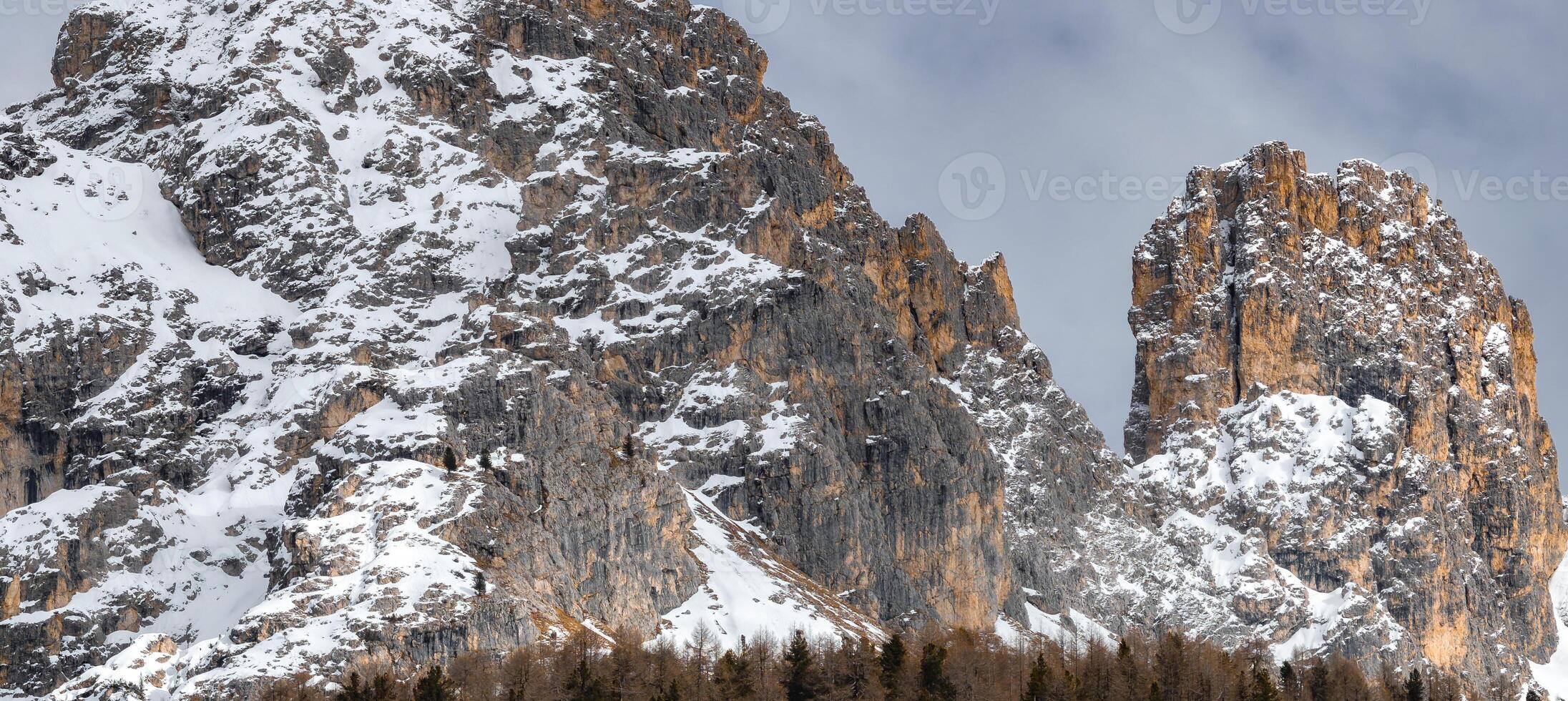 Towering rocky peaks of the Dolomites partially covered in snow, with evergreen trees at the base and an overcast sky creating a dramatic scene. photo