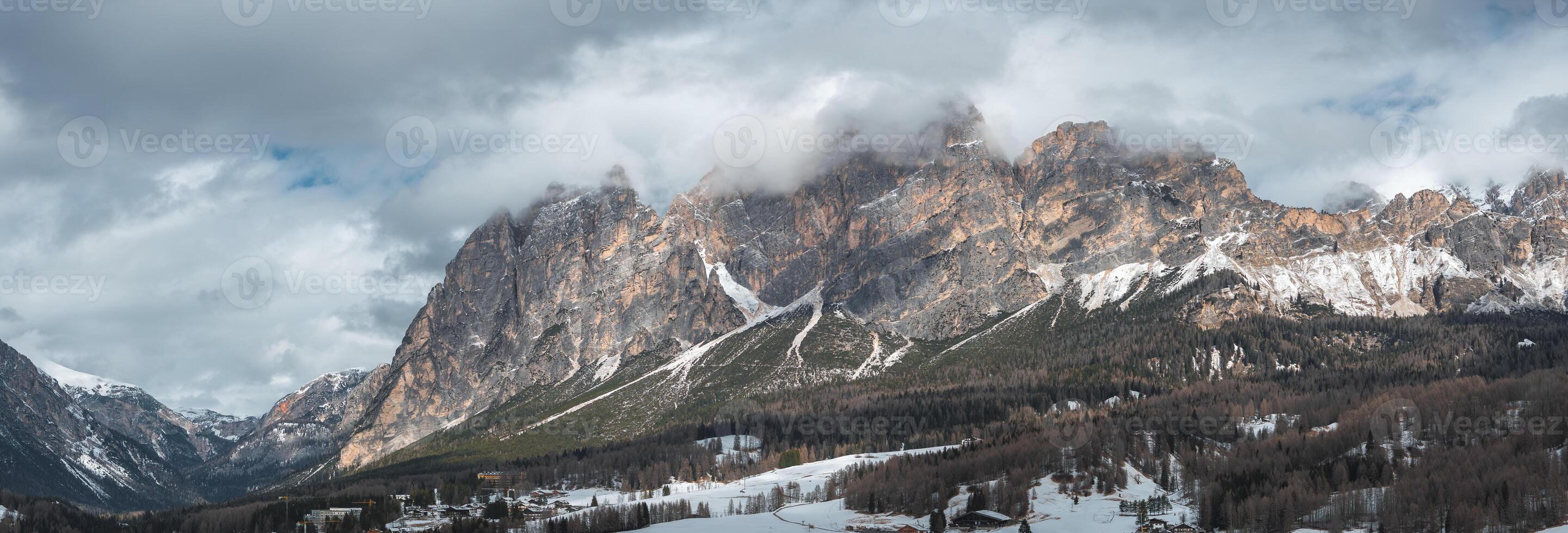 Aerial view of rocky Dolomite peaks with snow and clouds, a forested valley, ski slopes, and buildings near Cortina d'Ampezzo, Italy. photo