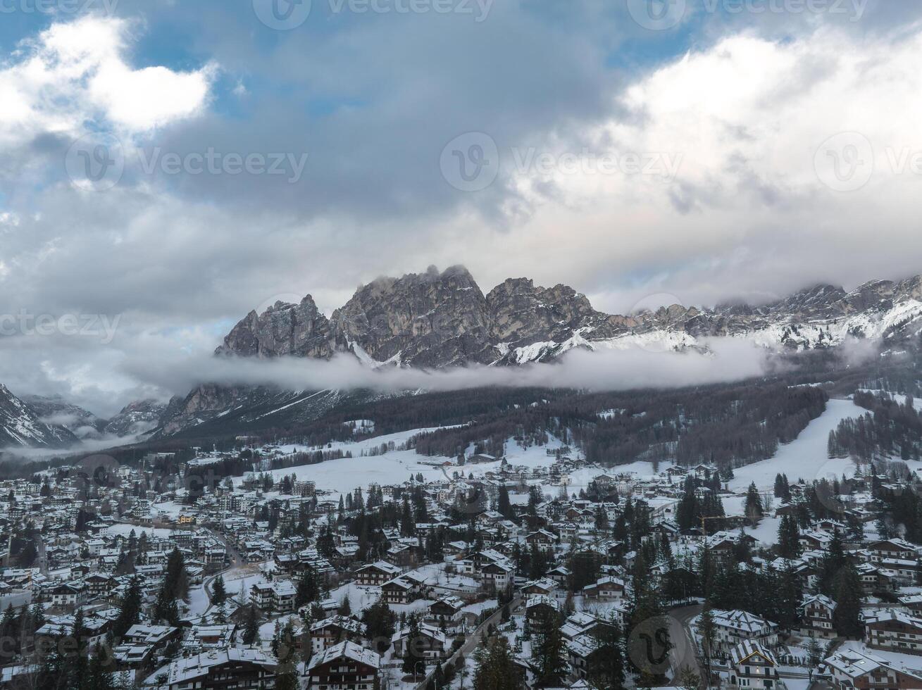 Aerial view of Cortina d'Ampezzo, Italy, with snow covered rooftops, alpine buildings, winding roads, and the Dolomites partially veiled by clouds. photo