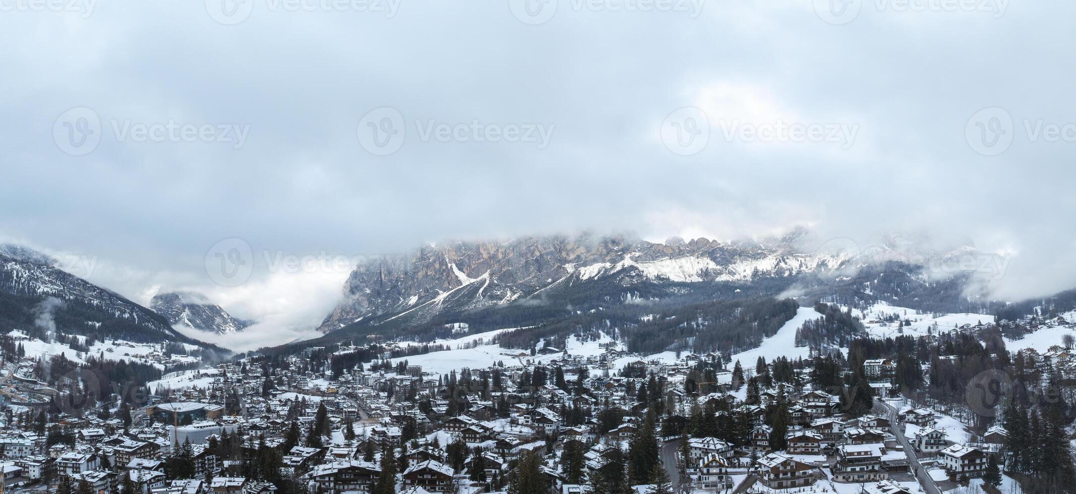 Aerial view of Cortina d'Ampezzo, Italy, with snow covered rooftops, alpine architecture, and the Dolomites partially shrouded in clouds. photo