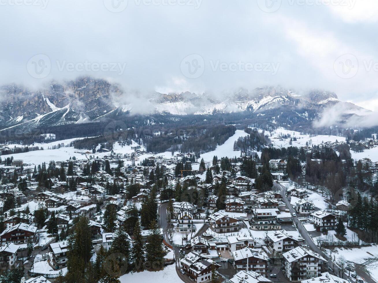 Aerial view of Cortina d'Ampezzo in Italy, featuring snow covered alpine architecture, dense forests, ski runs, and Dolomites peaks under cloudy skies. photo