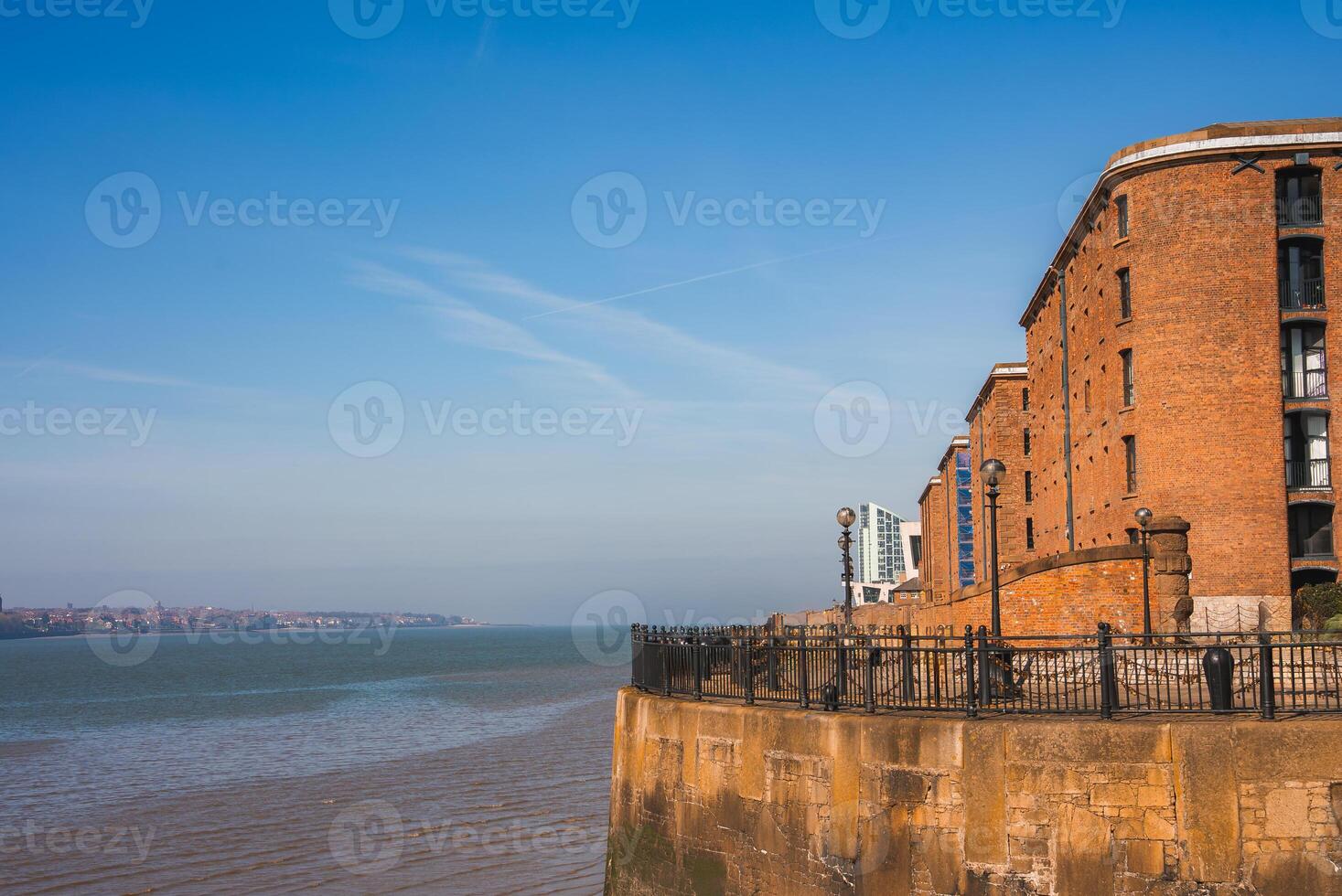 Historic Liverpool waterfront featuring a red brick Albert Dock building, curved design, iron railings, and the River Mersey under a clear blue sky. photo