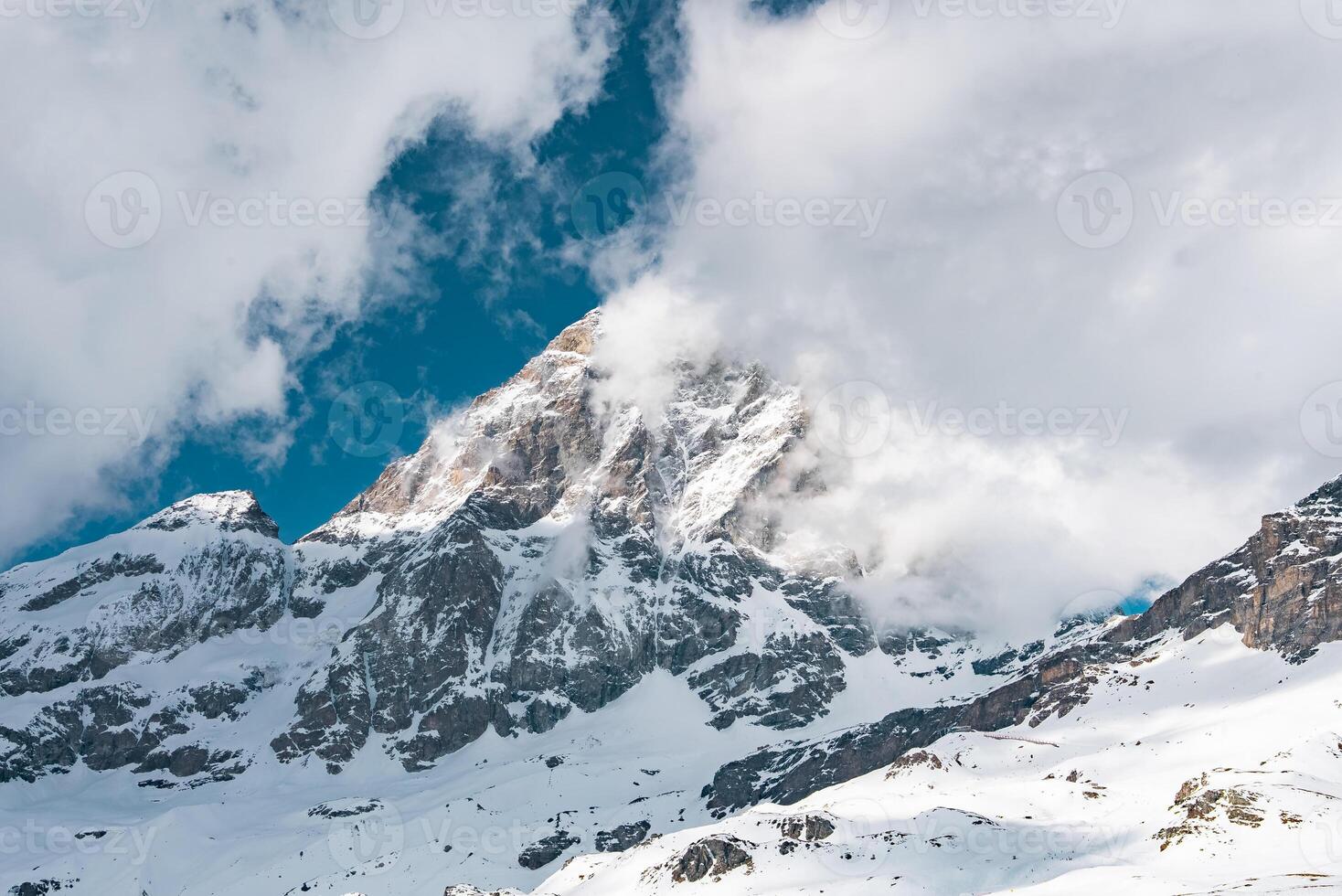 A snow covered mountain peak in Zermatt, Switzerland, partially shrouded in clouds. Sunlight and shadows highlight the rugged alpine terrain. photo