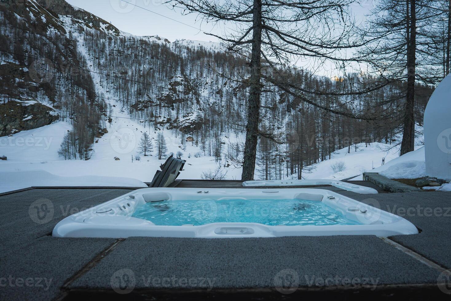 An outdoor hot tub on a wooden deck with steam rising, surrounded by snow covered slopes, sparse trees, and rugged mountain terrain in Zermatt, Switzerland. photo