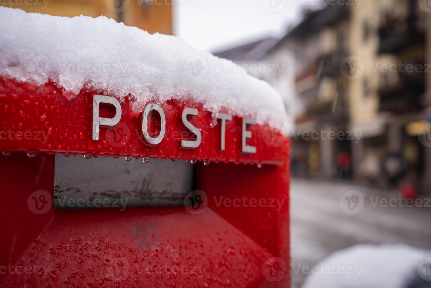 A red postal box labeled 'POSTE' is partially covered in snow, set against a blurred view of buildings with balconies in a snowy town, likely Cortina. photo
