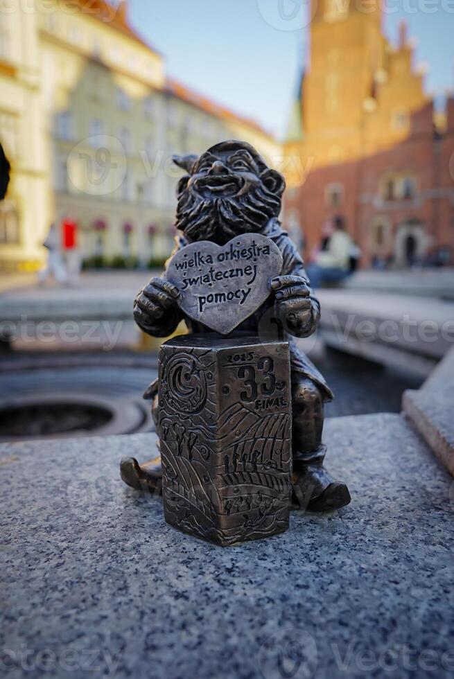 Small bronze gnome statue holding a heart shaped plaque, set near a fountain with Wroclaw's historic Market Square architecture in the background. photo