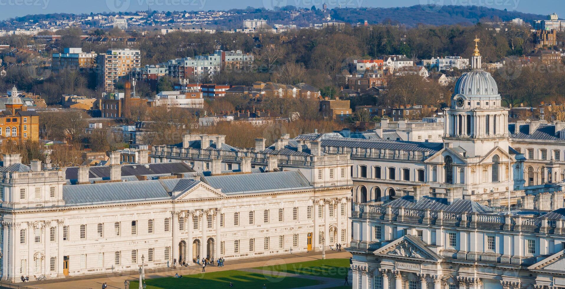 The Old Royal Naval College in Greenwich, London, features grand neoclassical design, domed structures, and a backdrop of residential areas and wooded hills. photo
