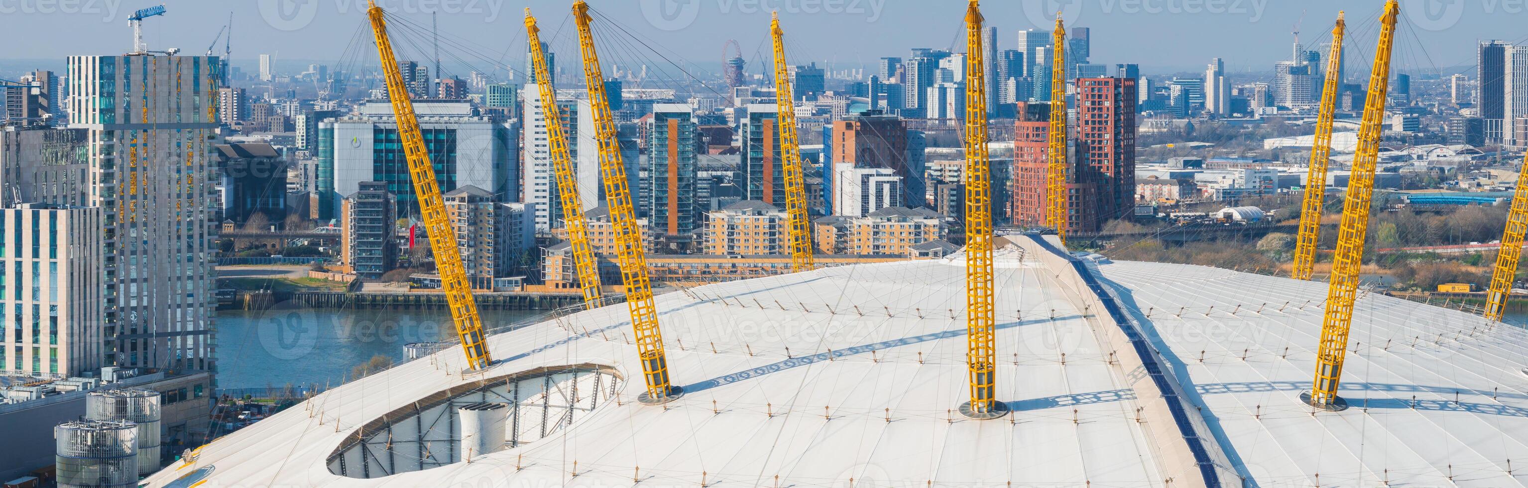 The O2 Arena's white dome and yellow masts stand out amid modern high rises, with the River Thames and Canary Wharf visible in the background. photo