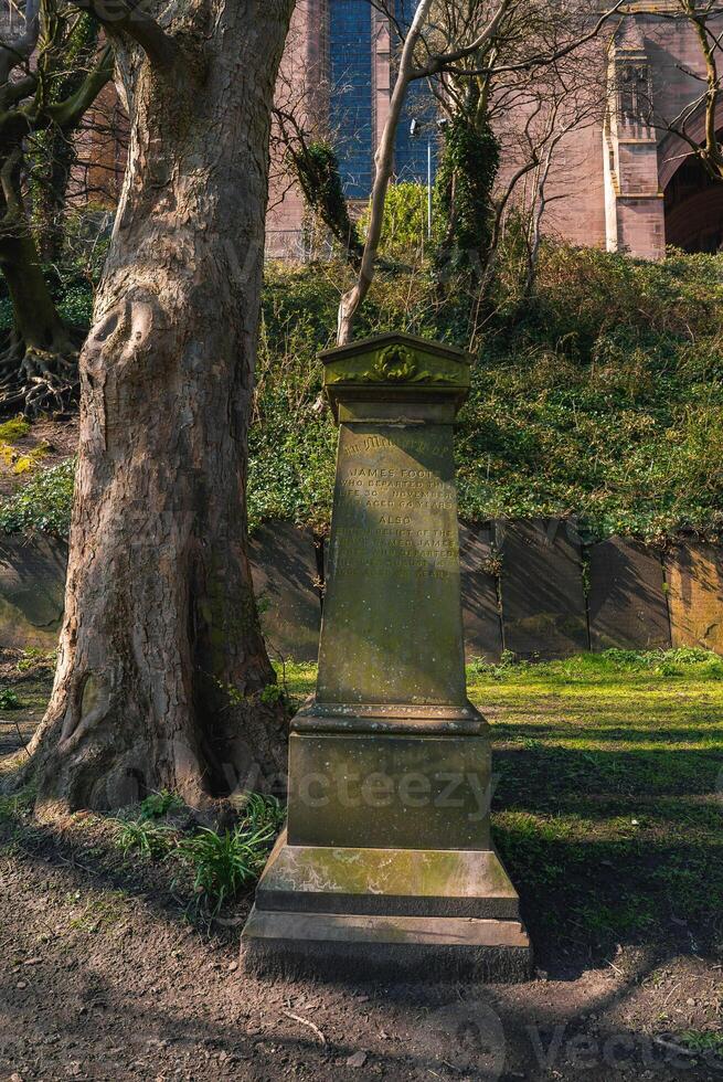 A weathered stone monument surrounded by greenery and a large tree, with Liverpool Cathedral's red sandstone facade visible in the background. photo