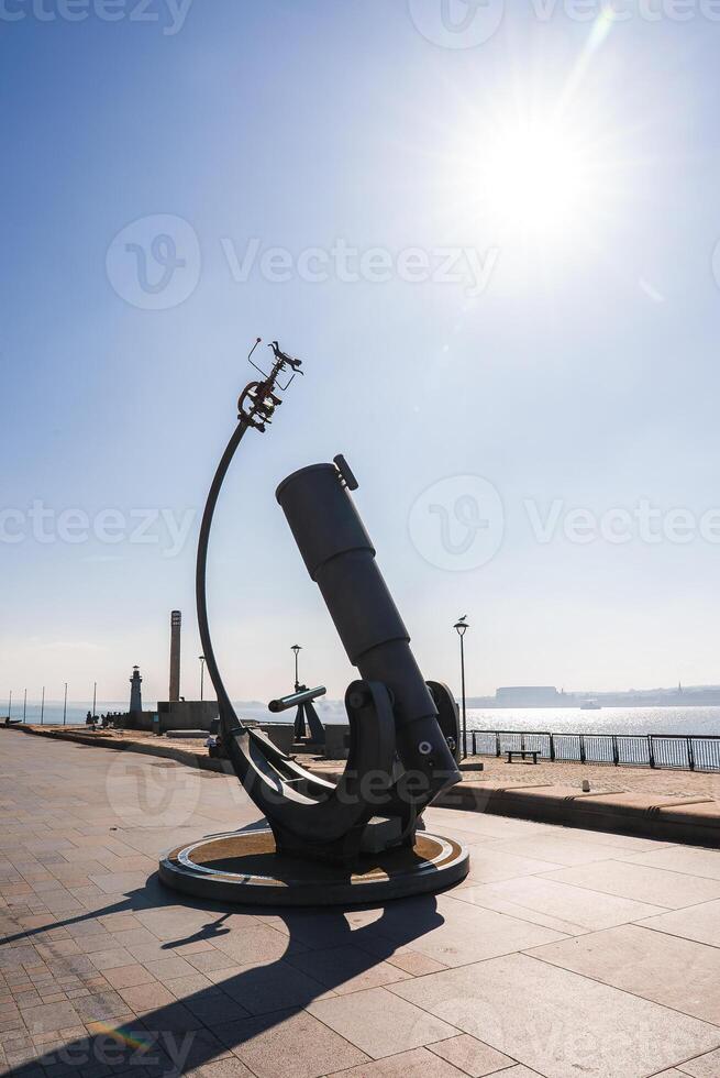 A large telescope like sculpture on a paved promenade in Liverpool, with the River Mersey, industrial structures, and a tall chimney in the background. photo