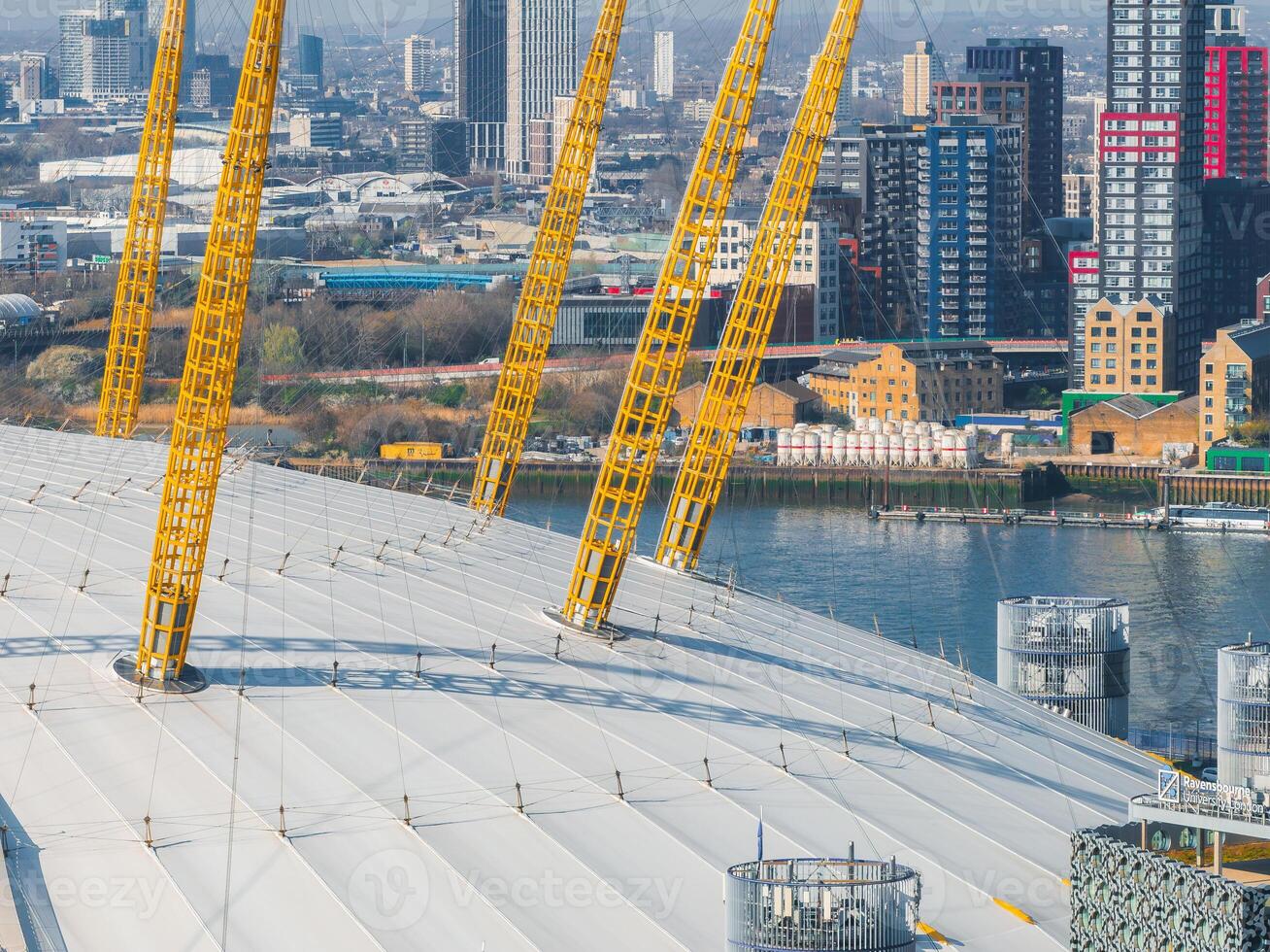 Close up aerial view of the O2 Arena in London with its white dome and yellow masts, the River Thames, and modern high rise buildings in the background. photo