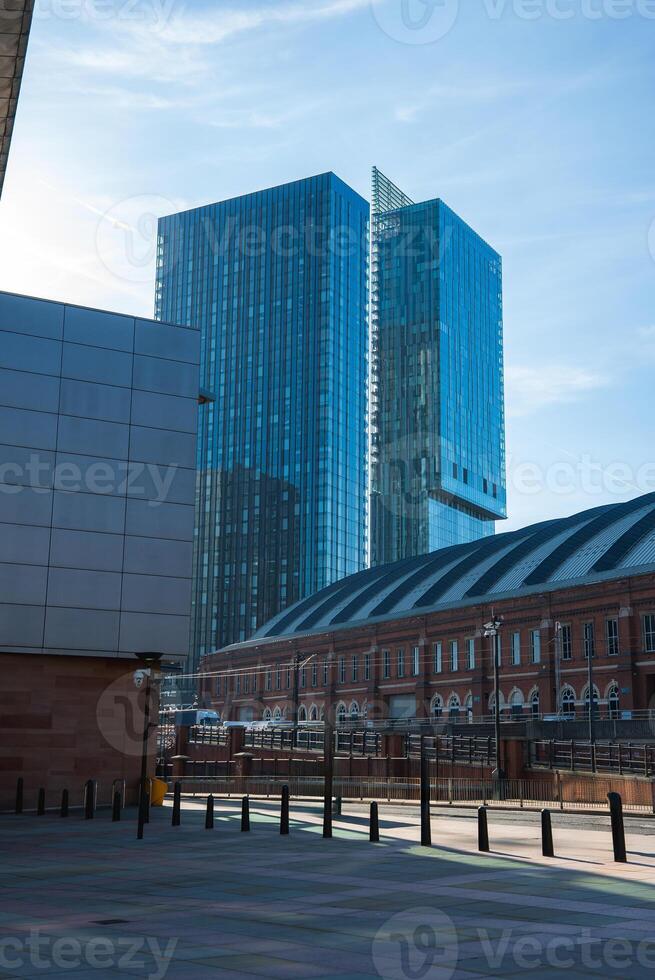 View of Manchester featuring Deansgate Square towers, a historic red brick building, a paved area with bollards, and a clear blue sky. photo