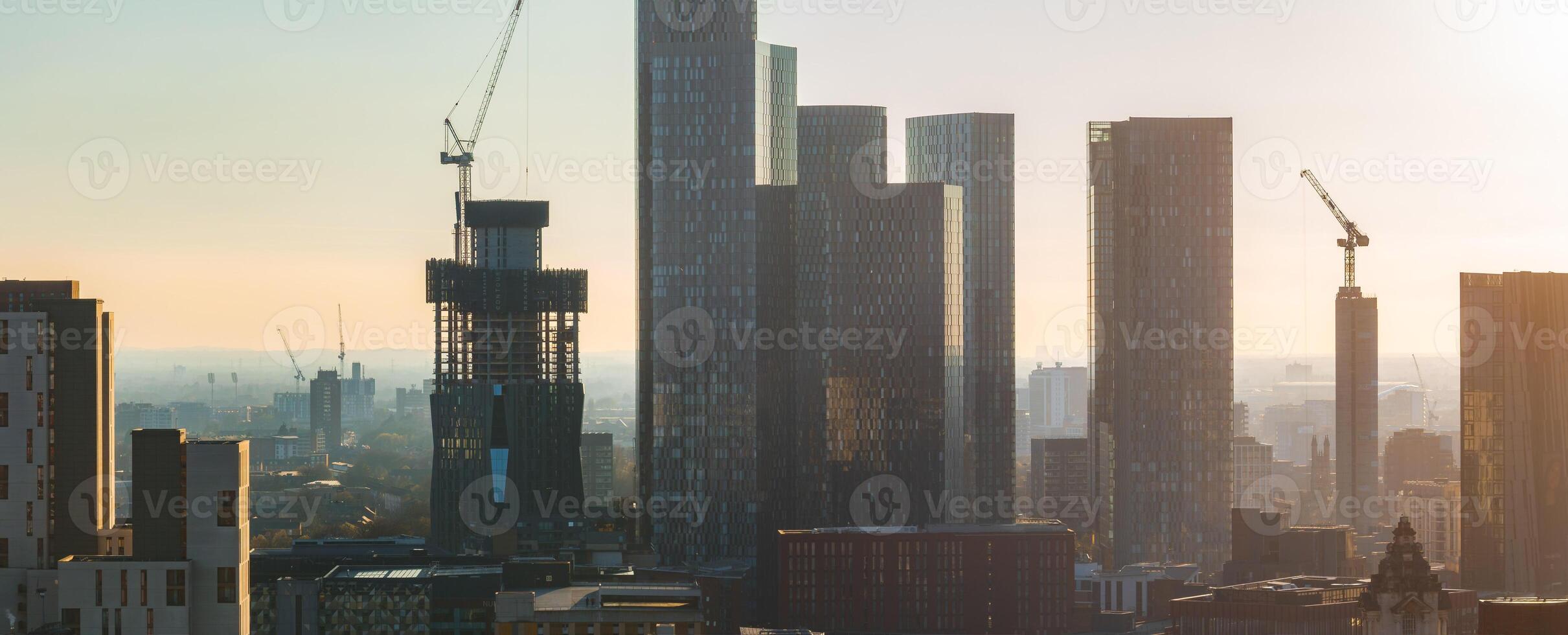 Aerial view of Manchester, UK, featuring modern skyscrapers, glass clad buildings, and a crane atop an unfinished high rise on a sunny day. photo