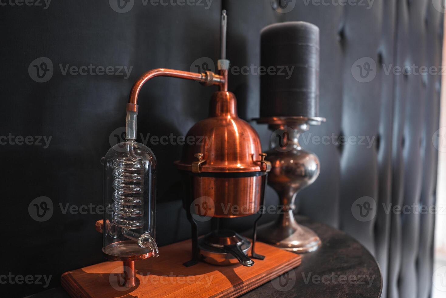 A vintage copper distillation apparatus with a glass condenser sits on a wooden base beside a black candle in a silver holder on tufted black leather. photo