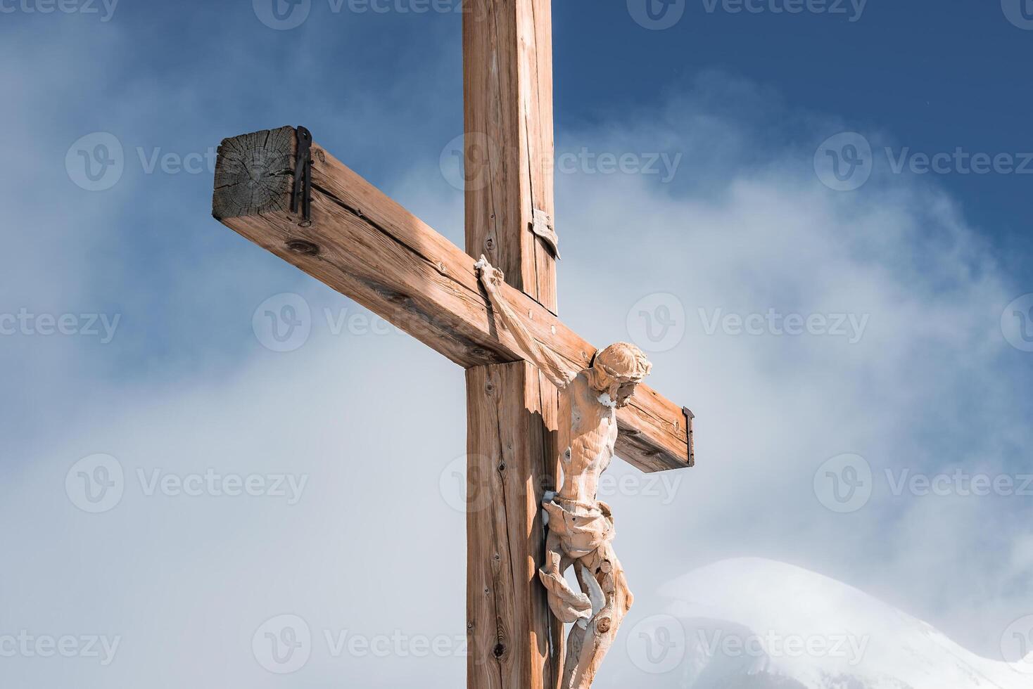 A detailed wooden crucifix of Jesus Christ stands against a clear blue sky, with snow covered mountain peaks and soft clouds in the Zermatt Alps. photo