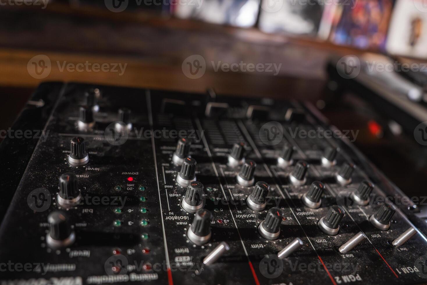 A professional audio mixer with knobs, sliders, and buttons on a wooden surface in a dimly lit room. Blurred album covers are visible in the background. photo