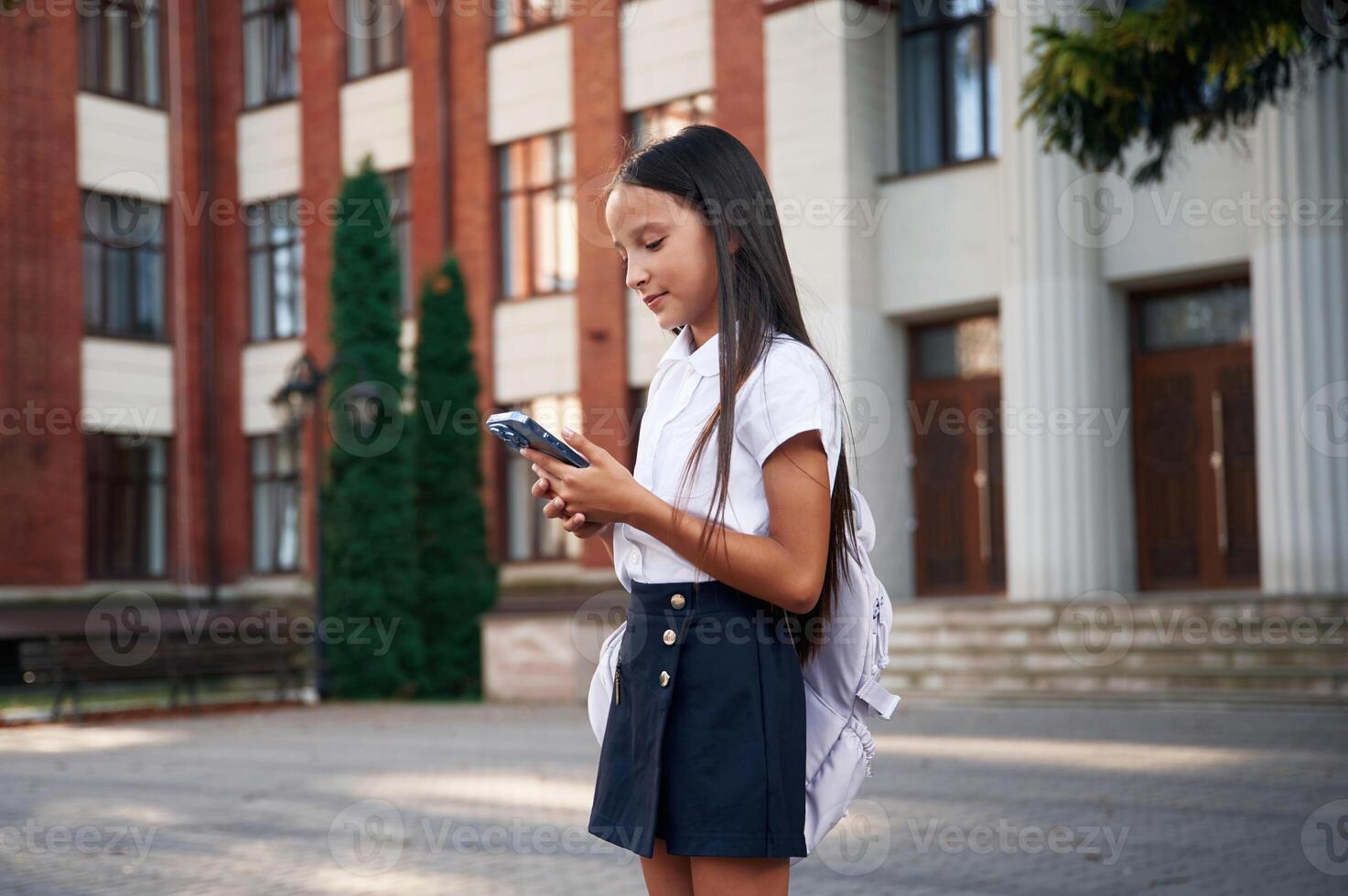 Wireless ,device, in hands, standing. School girl in uniform is outdoors near the building photo
