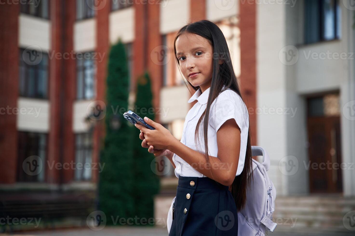 Holding smartphone. School girl, in ,uniform is outdoors near the building photo