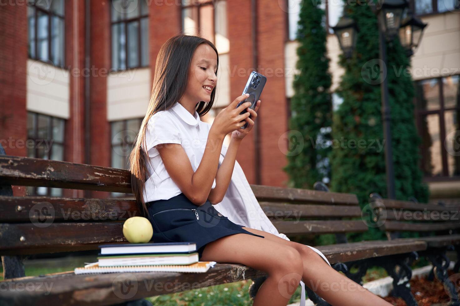 Sitting on the bench with smartphone. School girl in uniform is outdoors near the building photo