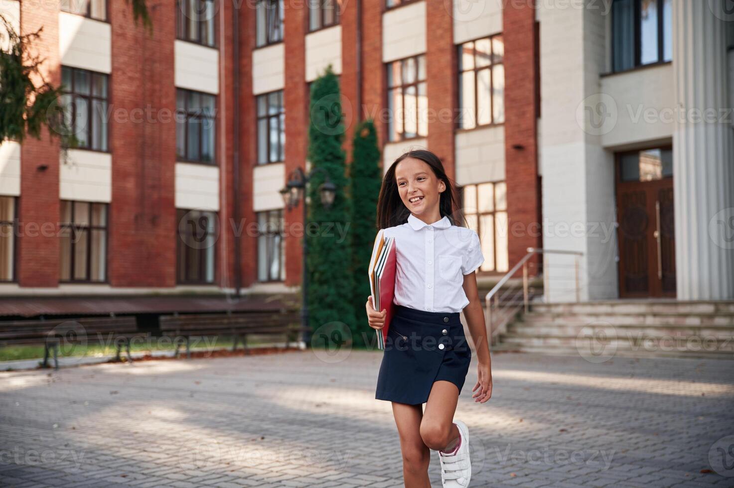 Standing, front view,, holding books., School girl in uniform is outdoors near the building photo