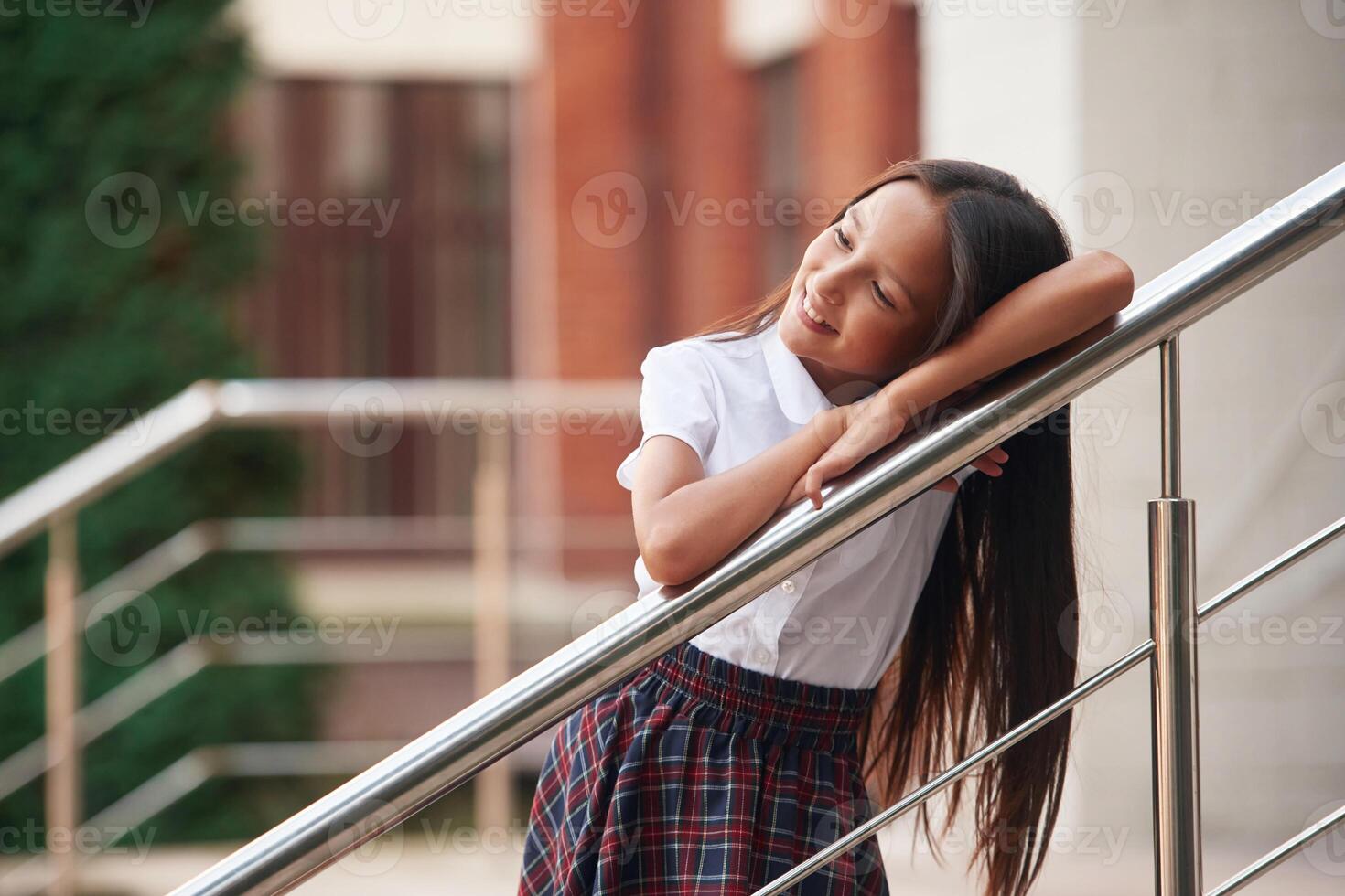 Leaning, on the ,railing. School girl in uniform is outdoors near the building photo