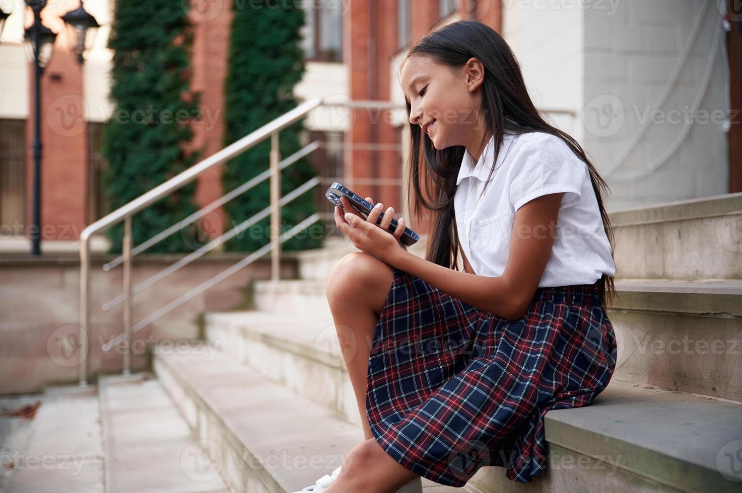 Sitting on the stairs,, using ,smartphone. School girl in uniform is outdoors near the building photo