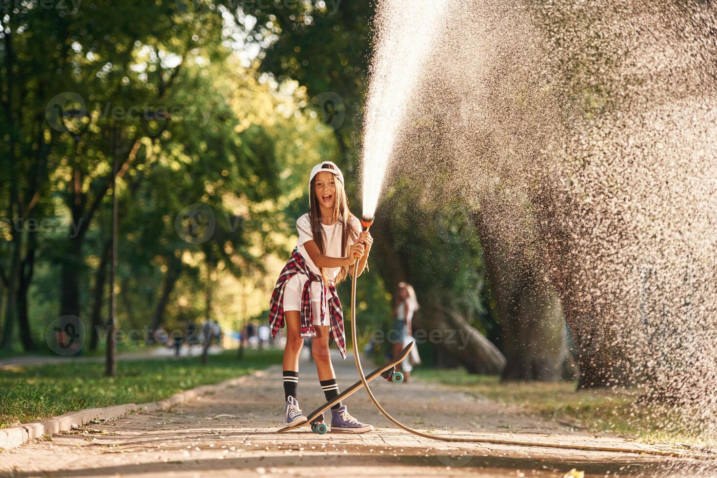 Plays with water as it pours from a hose. Happy little girl with skateboard outdoors photo