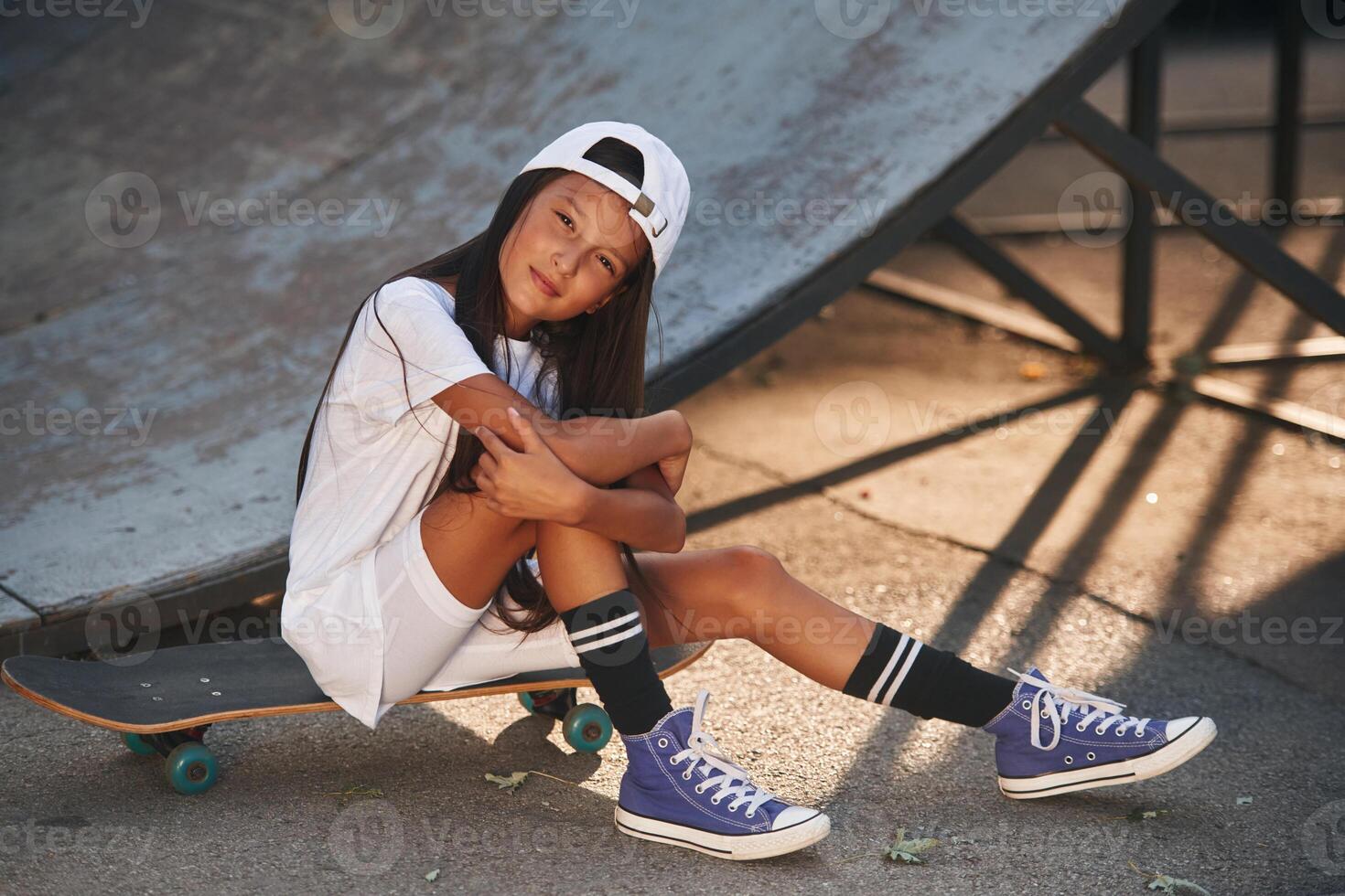 Ramp is behind. Girl is sitting. ,With skateboard outdoors photo