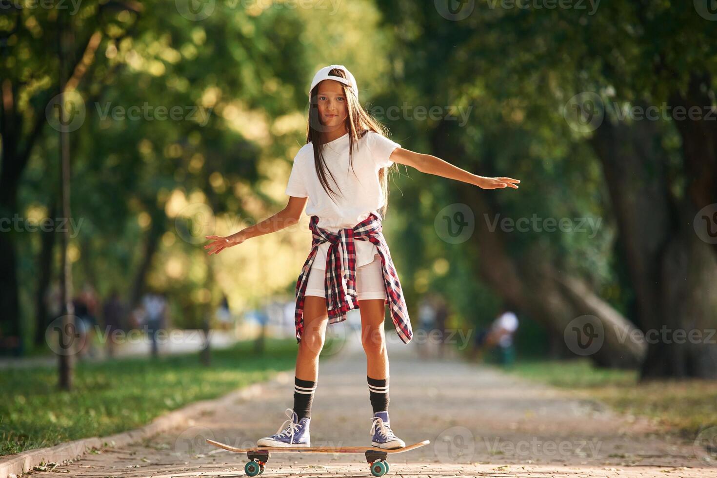 Green ,trees are in the background, on the path. Happy little girl with skateboard outdoors photo
