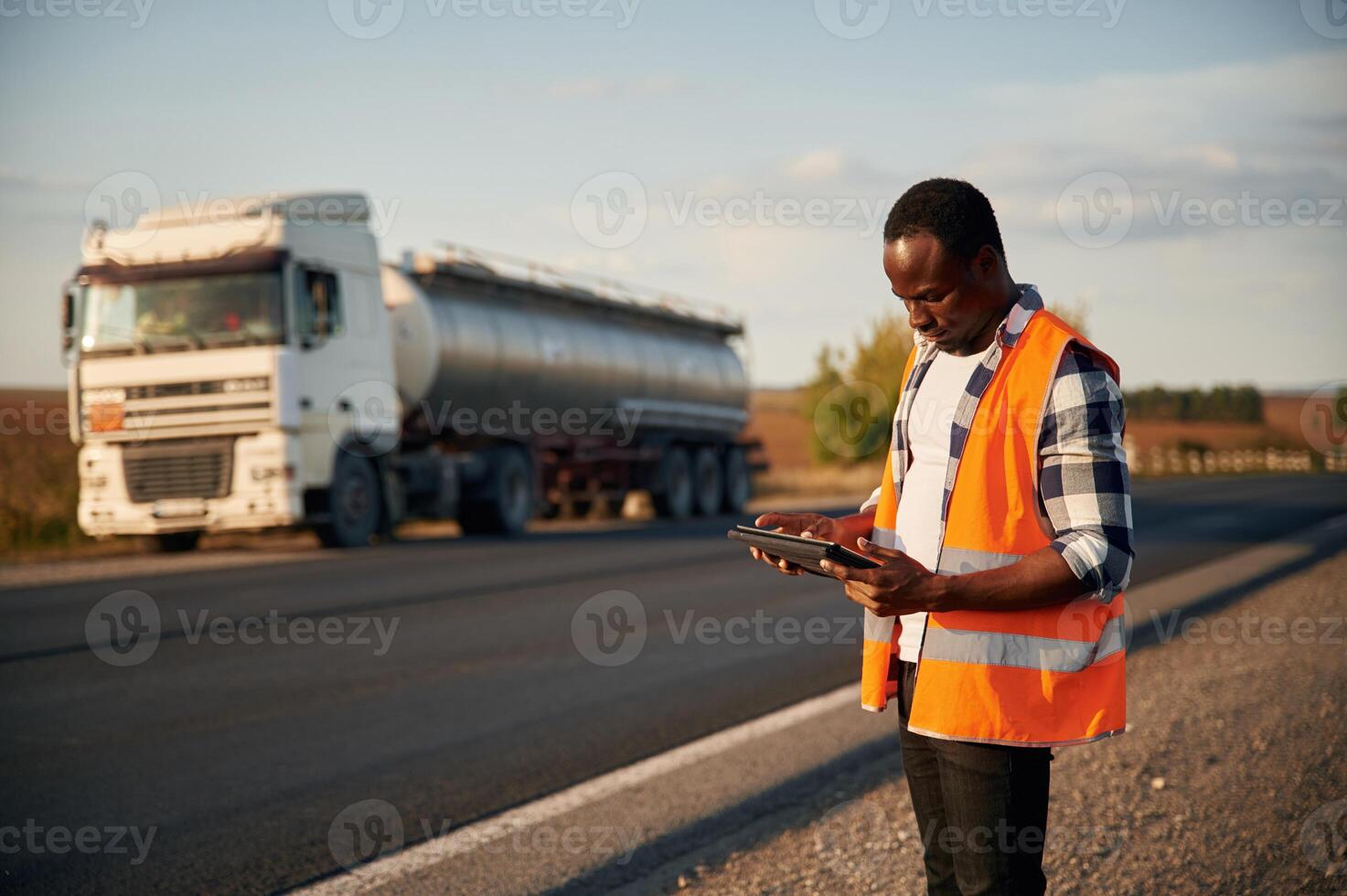 In ,orange colored uniform, ,with ,tablet. Black man is standing on the road with truck on it photo