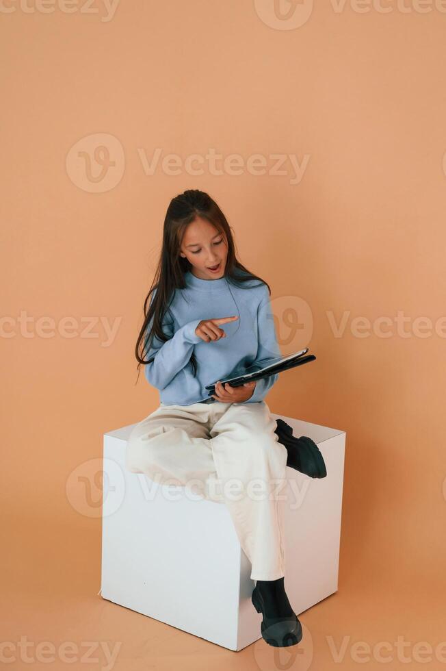 With tablet in hands. Sitting, on cube. Cute young girl is in the studio against background photo
