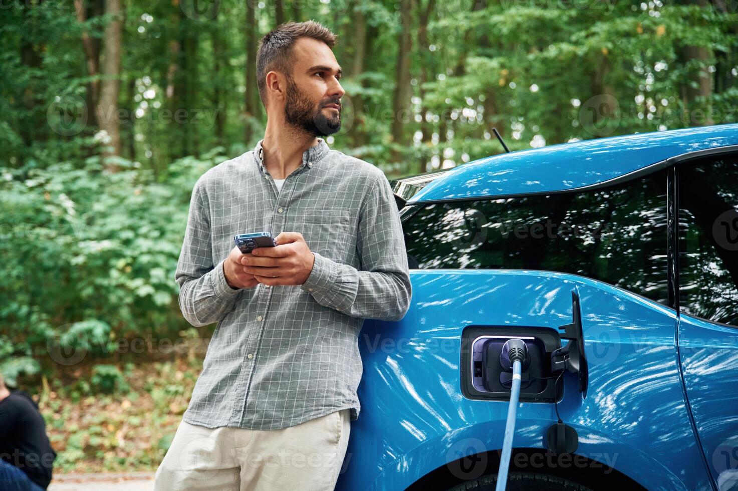 Trees behind.. Handsome ,,man is standing outdoors near electric automobile and waiting for charging photo