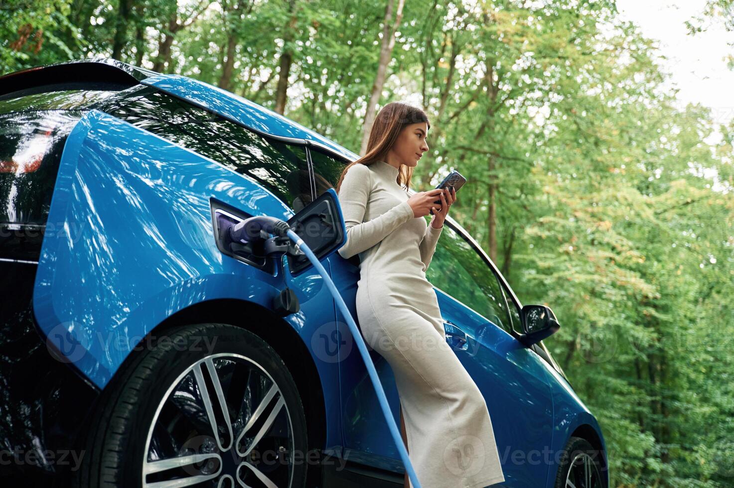 Leaning,,, using smartphone. Young woman is standing outdoors near electric automobile that is charging photo