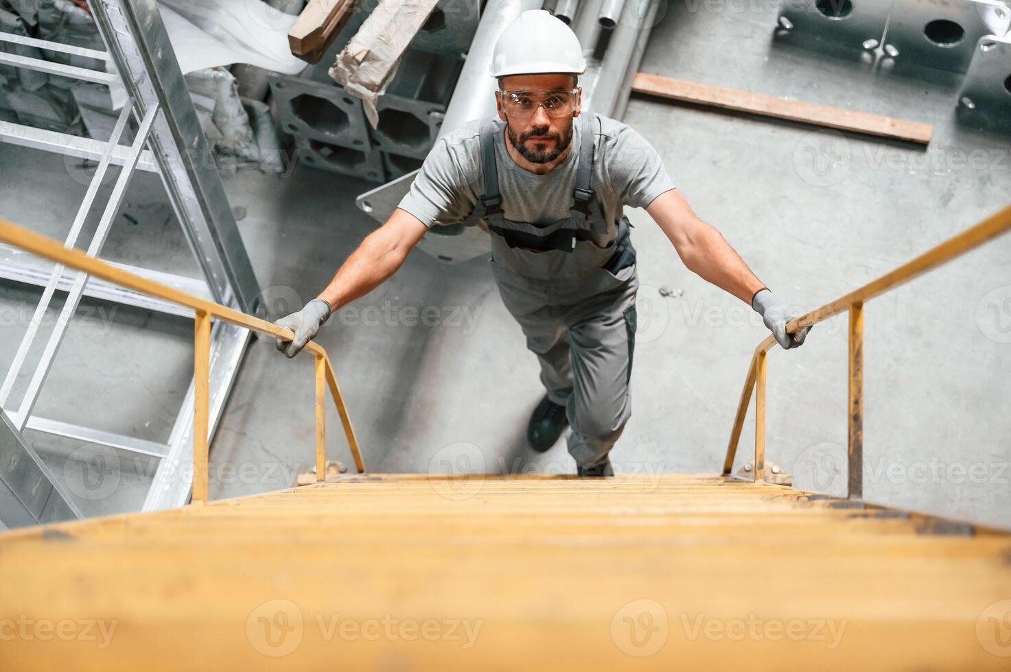 Going,, up on the ladder, top view. Young factory worker in grey uniform photo