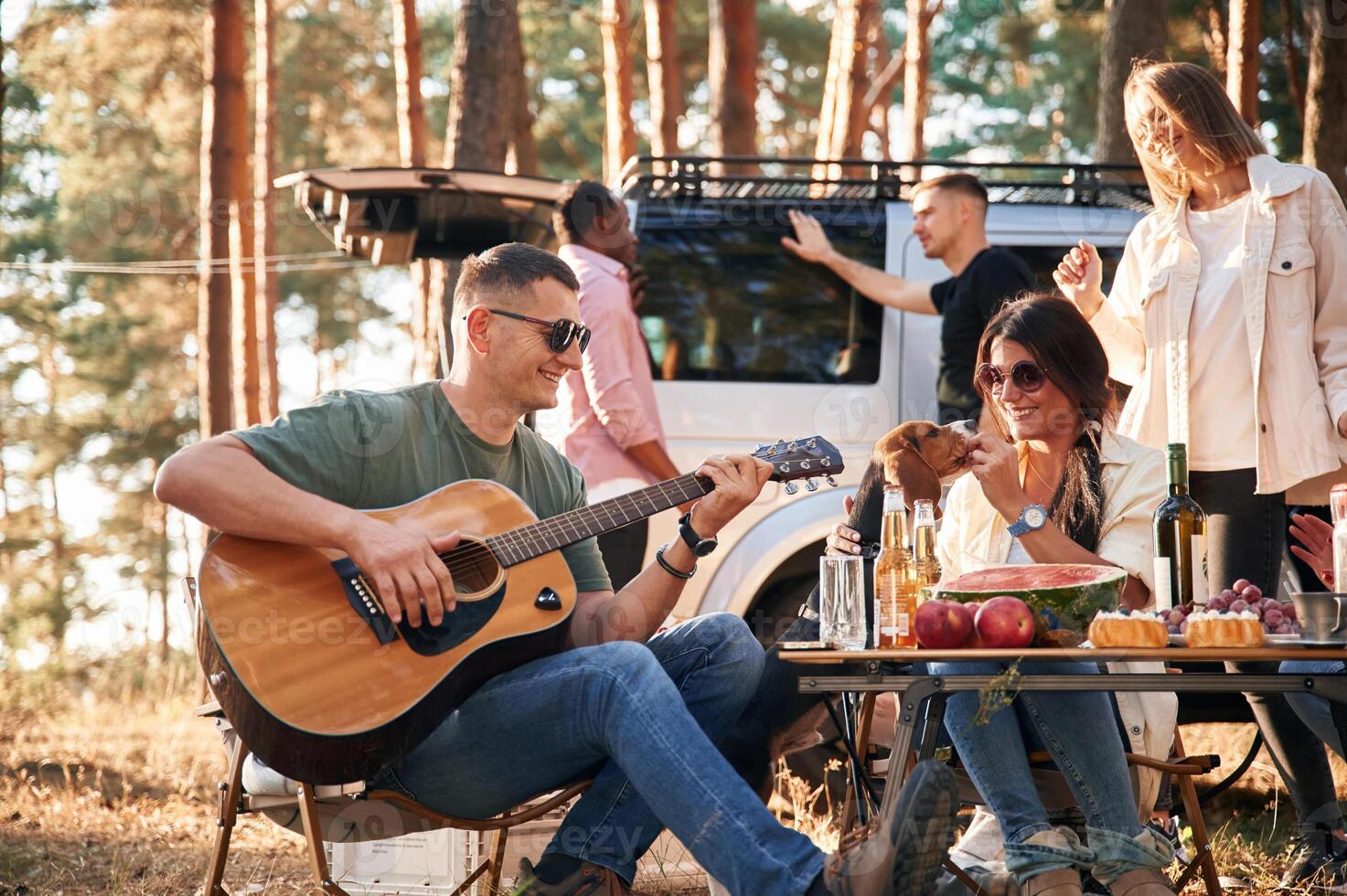 Man is playing guitar, by table. Group of friends are together in the forest photo