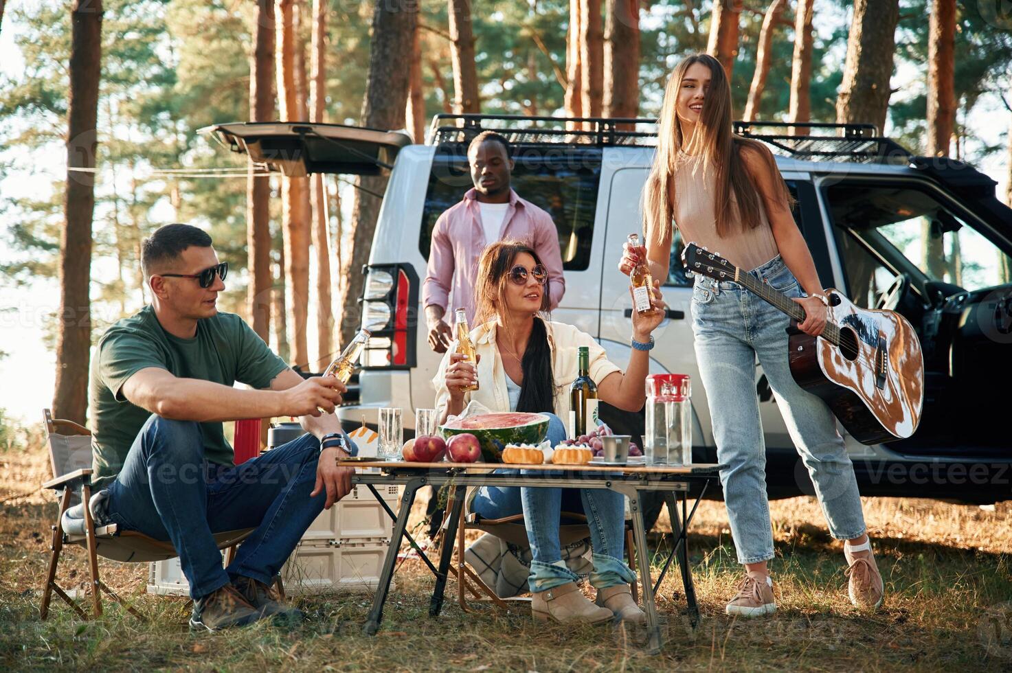 Sitting, by the table with food, against car. Group of friends are together in the forest photo