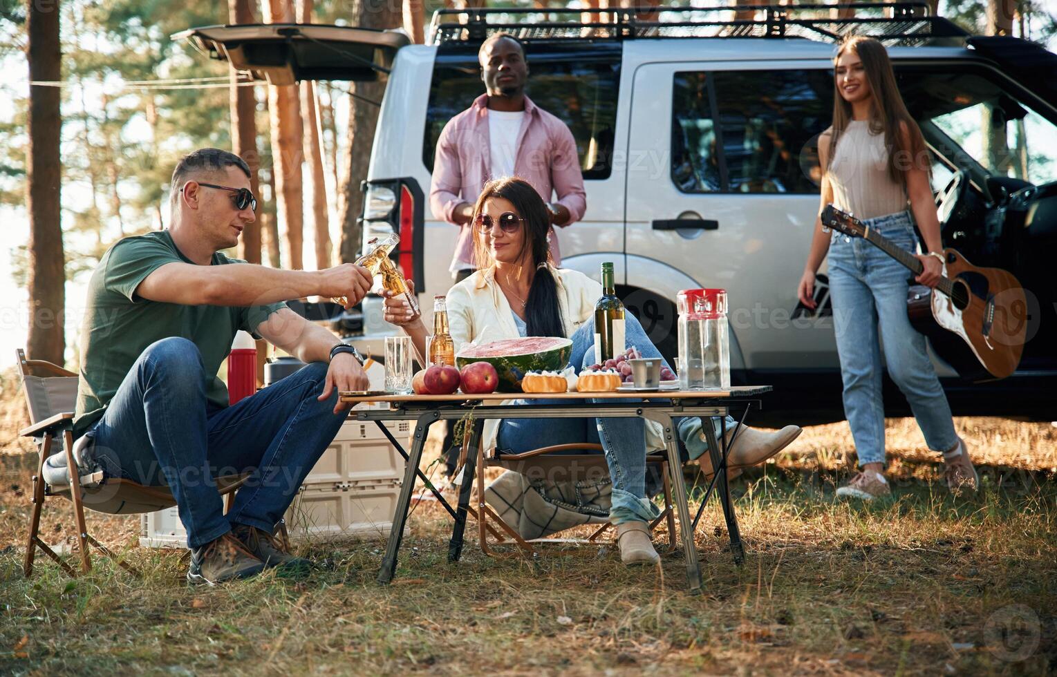 Sitting ,by the ,table with food, against car. Group of friends are together in the forest photo