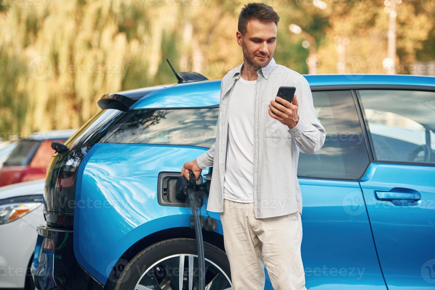 Wire in ,socket, holding ,smartphone.. Man with blue electric car on the charge station photo
