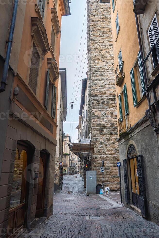A quiet cobblestone street in Bergamo's old town, lined with warm toned buildings, green shutters, a stone tower, utility wires, and a streetlight. photo