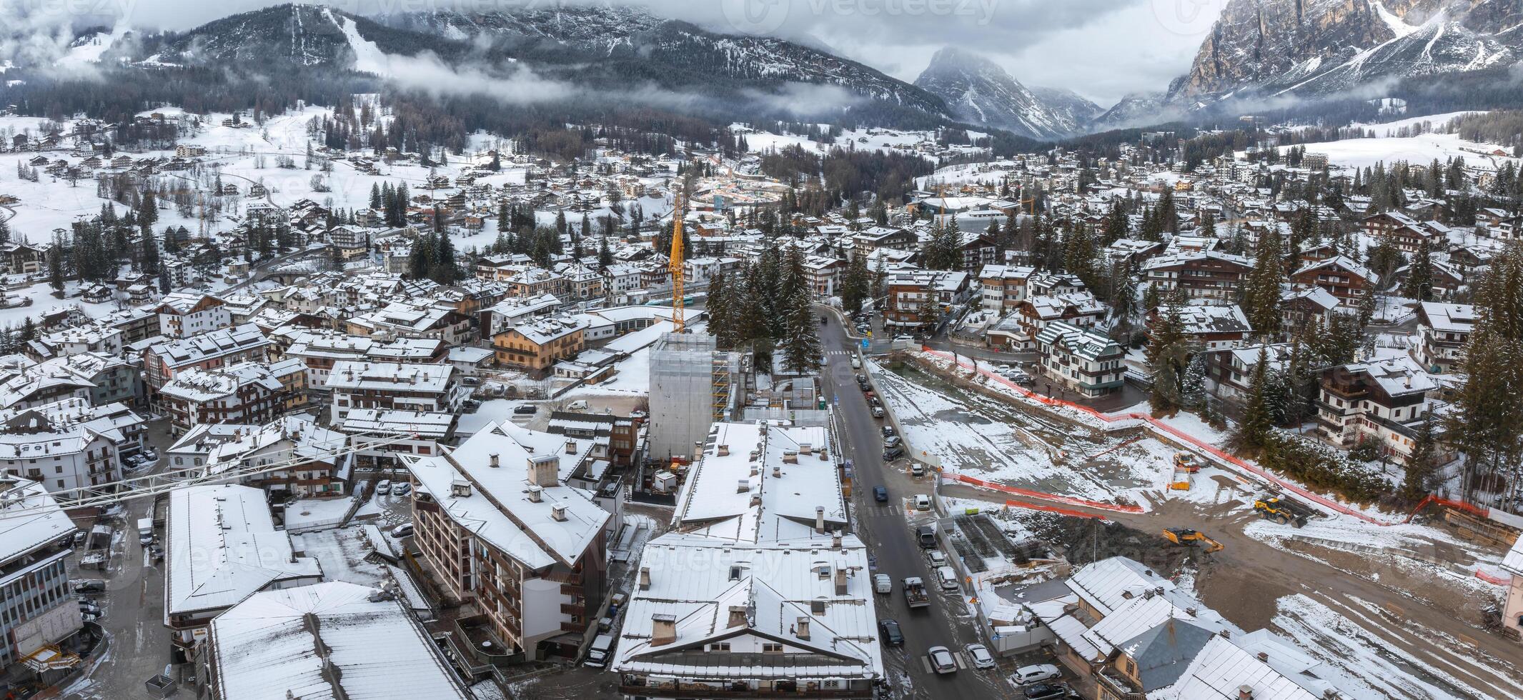 Aerial view of Cortina d'Ampezzo in Italy, showing snow covered rooftops, alpine architecture, a construction site, and dramatic mountain peaks. photo