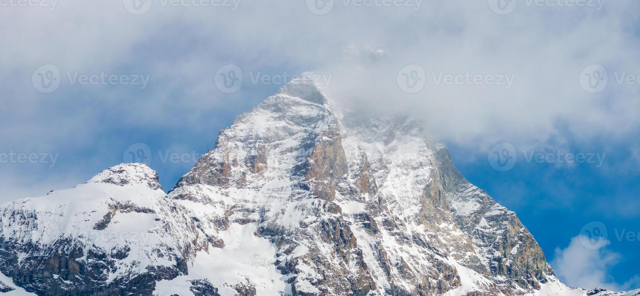 The Matterhorn mountain in the Alps features rugged, snow covered slopes, a triangular peak, and wisps of clouds against a clear blue sky. photo