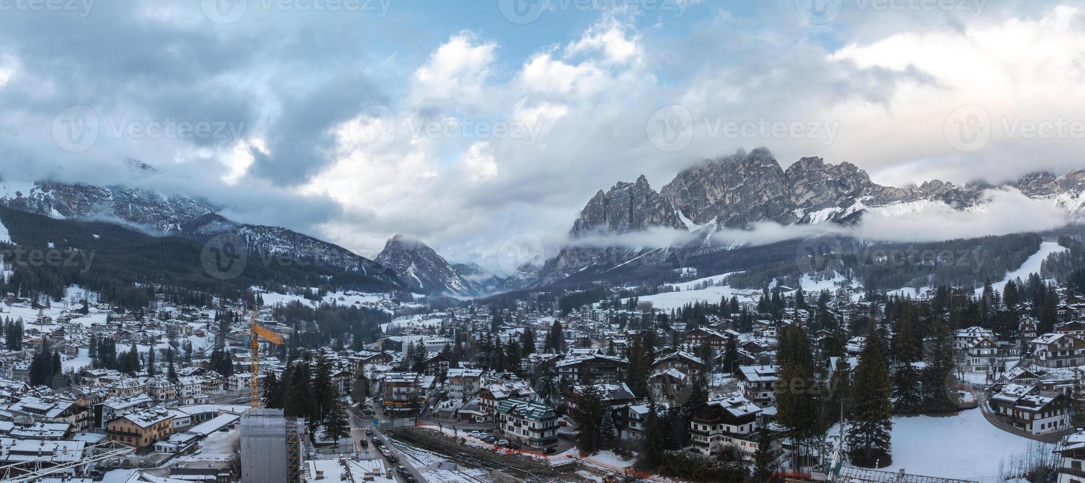 Aerial view of Cortina d'Ampezzo, Italy, showing snow covered rooftops, alpine buildings, forested valleys, mountain peaks, and construction activity. photo