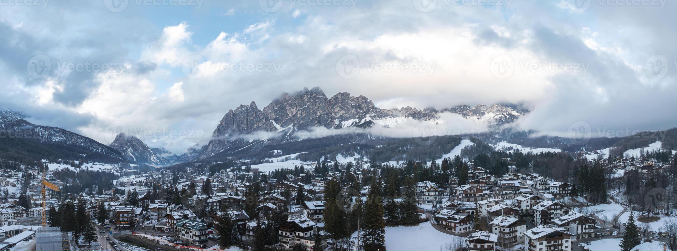 Aerial view of Cortina d'Ampezzo, Italy, showing snow covered rooftops, evergreen trees, Dolomite peaks, and a crane amid Winter Olympics preparations. photo